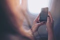 Woman holding and listening to music with smart phone , sitting next to an airplane window with clouds and sky background Royalty Free Stock Photo