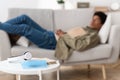 Blurred Black Teenage Guy Lying On Sofa At Home, Selective Focus Of Medical Mask, Book And Headphones. Copy Space