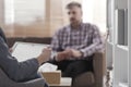 Blurred view of a single man sitting on a sofa during a psychotherapy session with a side view of the adviser holding a tablet in