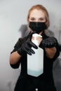 Blurred young woman in black wear and gloves and surgical mask with blue alcohol in her hands in bottle in focus.