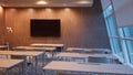 Blurry classroom scene focusing on empty desks with a large display screen on the wall illuminated by indoor lighting and Royalty Free Stock Photo