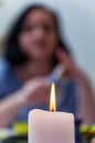 A blurry silhouette of a brunette sitting at a table. In the foreground is a lighted white candle. The concept of loneliness,