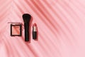 Blush, makeup brush, lipstick on a pink background with a shadow from a palm leaf top view. Summer makeup concept, women`s beauty