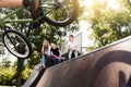 Bmx bicycle rider making tricks and sports kids with skateboard and penny boards are sitting and chatting on background