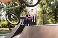 Bmx bicycle rider making tricks and sports kids with skateboard and penny boards are sitting and chatting on background