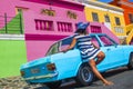 A beautiful African woman in a blue and white striped dress in front of a vintage Ford Cortina and traditional homes of Bo-Kaap, C