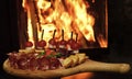 Board with snacks on wooden table, fire on background. Cold appetizers with tomatoes, sausage, salami, ham, arugula Royalty Free Stock Photo