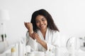 Body nourishing products. Pretty black lady in bathrobe applying moisturizing lotion or cream on body after shower Royalty Free Stock Photo