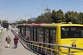 Two persons walking into a Transmilenio station with a big red and yellow bus near to them