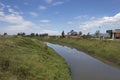 Bogota river crossing green country field with near modern buildings at sides and blue sky at background