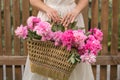 Boho girl holding pink peonies in straw basket. Stylish hipster woman in bohemian floral dress gathering peony flowers on wooden