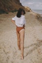 Boho girl in white shirt running on sunny beach, back view. Carefree stylish woman in swimsuit and shirt relaxing on seashore. Royalty Free Stock Photo