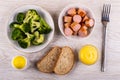 Boiled broccoli in plate, pieces sausages, salt, bread, mayonnaise, fork on table. Top view