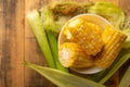 Boiled corn cob in a plate with butter, on wooden textures and green stalks of corn, tasty homemade food, for a picnic, with space Royalty Free Stock Photo