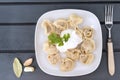 Boiled dumplings with sour cream and herbs on a white plate, fork, garlic, Bay leaf on a wooden gray background, top view