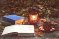 The book, lamp and a cup of hot coffee on the old wooden table in a forest. Fallen yellow maple leaves.