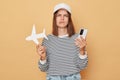 Booking an economy flight. Flying around the world. Unhappy woman wearing baseball cap and striped shirt holding white paper plane