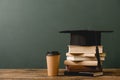 Books, academic cap and disposable cup on wooden surface