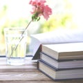 Books are ready for reading lying on a wooden windowsill next to a glass with clear water in which there is a pink geranium flower