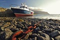 Boot moored at a jetty with pebble beach and rusty chain leading towards the shoreline in the foreground Royalty Free Stock Photo