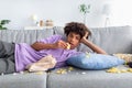 Bored Afro teenager lying on sofa with scattered snacks, eating chips, watching dull show or movie on TV at home