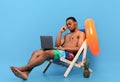 Bored black guy feeling unhappy, having to work online during his summer vacation, sitting in lounge chair with laptop