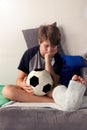 Bored boy with a broken leg sitting on the couch and holding a football ball.