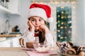 Bored cute little girl kid in Santa Claus hat sitting on the decorated for winter holidays modern kitchen with cocoa Royalty Free Stock Photo