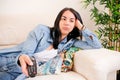 Woman who is lying on her sofa, feeling bored and uninterested while zapping TV channels with her remote control