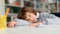 Bored european little schoolboy looking away, leaning on table in library or classroom during uninterested lesson