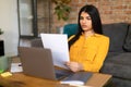 Bored female teenager sitting at table with papers, using laptop to write coursework, studying online from home