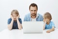 Bored indifferent son sitting with sibling and father at table, leaning head on hands while looking at laptop screen