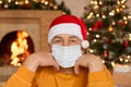 Bored mature male sitting in living room with New Year decoration, keeping fists under chin, wearing medical safety mask for