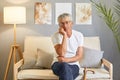 Bored sad upset grey-haired mature man wearing casual white T-shirt sitting on sofa at home interior felling boring holding chin Royalty Free Stock Photo