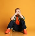 Bored schoolboy teenager, regular guy in checkered shirt, orange t-shirt and jeans sits holding fists at his cheeks Royalty Free Stock Photo