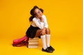 Bored Schoolgirl Sitting On Stack Of Books Over Yellow Background