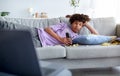 Bored teen guy lying on sofa with scattered snacks, watching movie on laptop at home