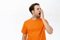 Bored or tired adult man yawning, cover his opened mouth with hand, stand in orange tshirt over white background Royalty Free Stock Photo