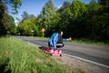 Bored of waiting to hitchhike, a teenager waits at the side of a road leading through a leafy forest on a sunny day. Royalty Free Stock Photo