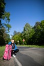 Bored of waiting to hitchhike, a teenager waits at the side of a road leading through a leafy forest on a sunny day. Royalty Free Stock Photo