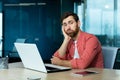 Boring office work. A young man sits in the office at a table with a laptop, leans on his arm and is bored, tired, looks