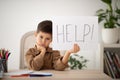 Boring tired sad small european boy sits at table shows paper with inscription help in school room interior Royalty Free Stock Photo