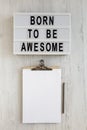 `Born to be awesome` words on a modern board, clipboard with blank sheet of paper on a white wooden background, top view. Overhe