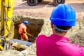 Boss oversees, controlling worker who is using shovel to set up right measures of square trench