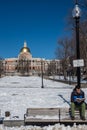 Boston, MA, USA, February, 8, 2016: A young man perches on the back of outdoor bench reading a book on a chilly snow covered park