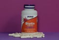 Bottle and capsules of vitamins Biotin 500 mcg manufactured by Now Foods on a purple background