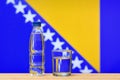 A bottle of clean drinking water and a glass stand on the table against the background of the flag of Bosnia and Herzegovina. Royalty Free Stock Photo