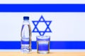 A bottle of clean drinking water and a glass stand on the table against the background of the flag of Israel.