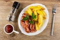 Condiment, ketchup, white dish with slices of fried potatoes, parsley, sausage, fork on wooden table. Top view