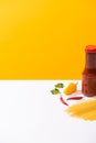 Bottle of tasty ketchup with peppers and raw spaghetti on white surface on yellow background